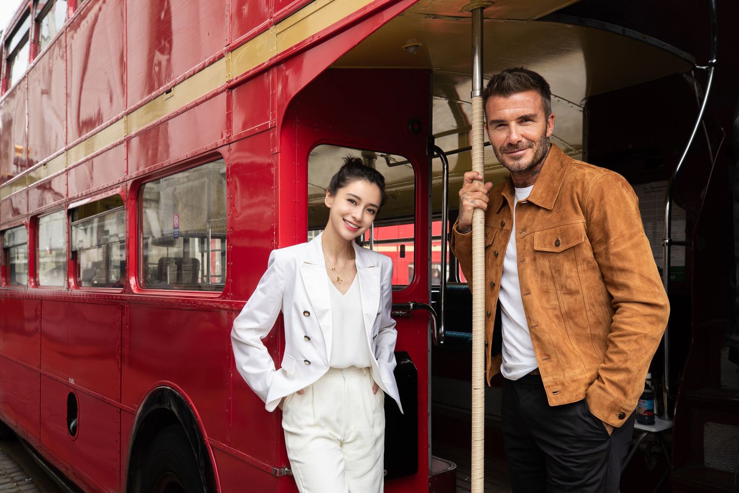 A stylish couple poses beside a classic red London double-decker bus on a city street.