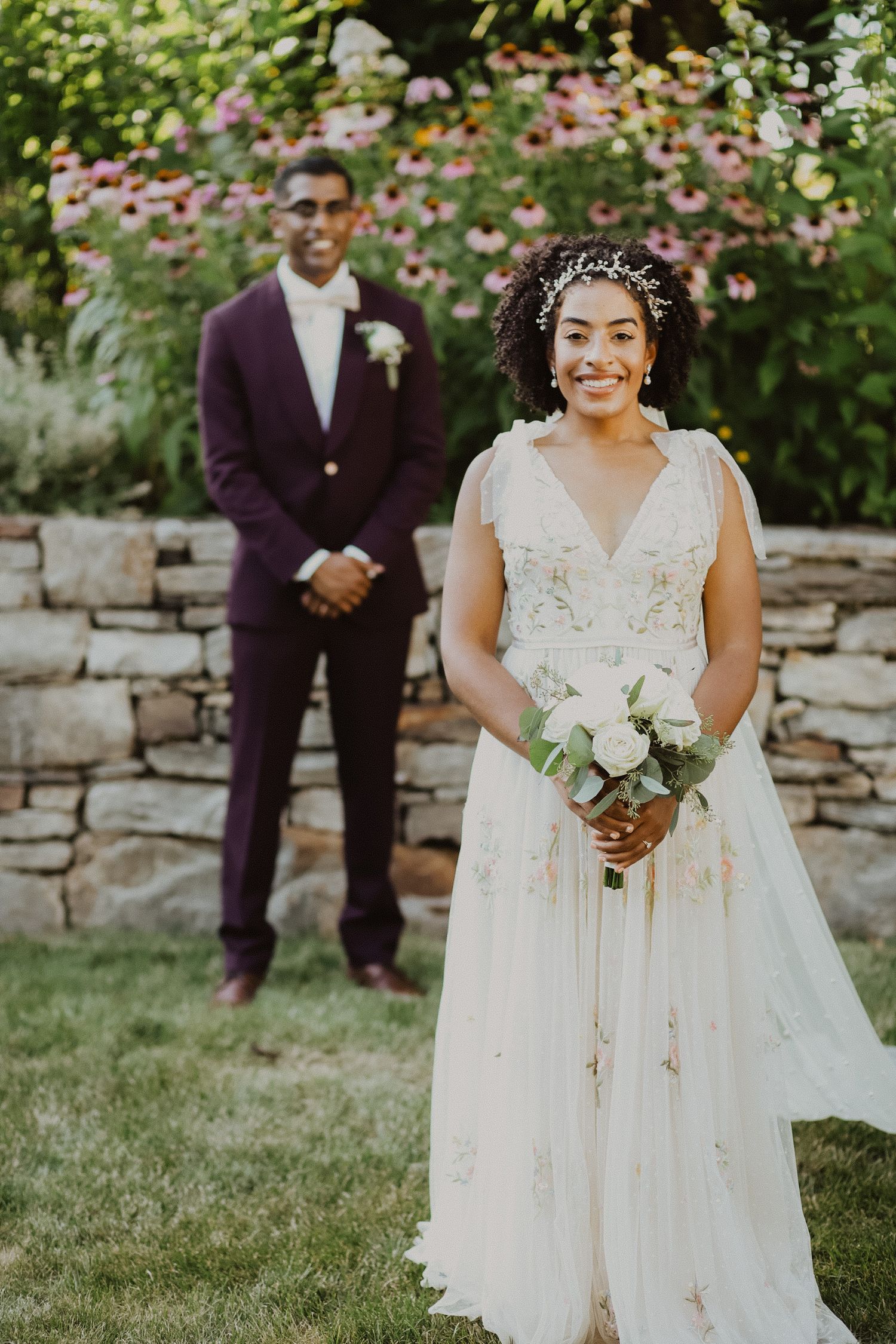 Bride in elegant lace dress poses in garden with groom standing in background.