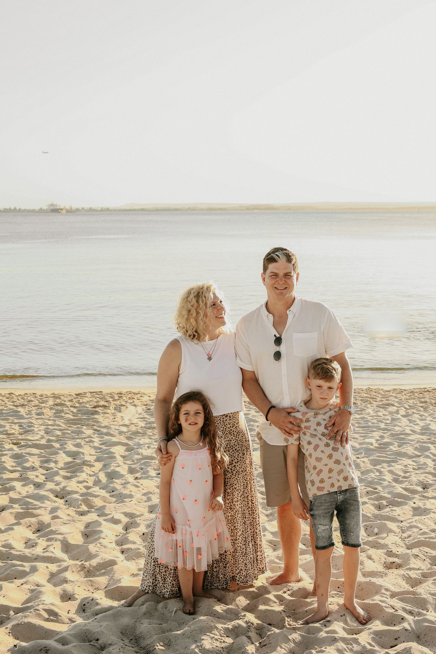 Happy family of four posing together on a sandy beach with calm ocean water in the background.