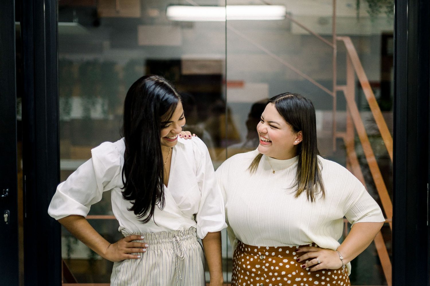 Two women laughing together in casual white tops against modern interior.