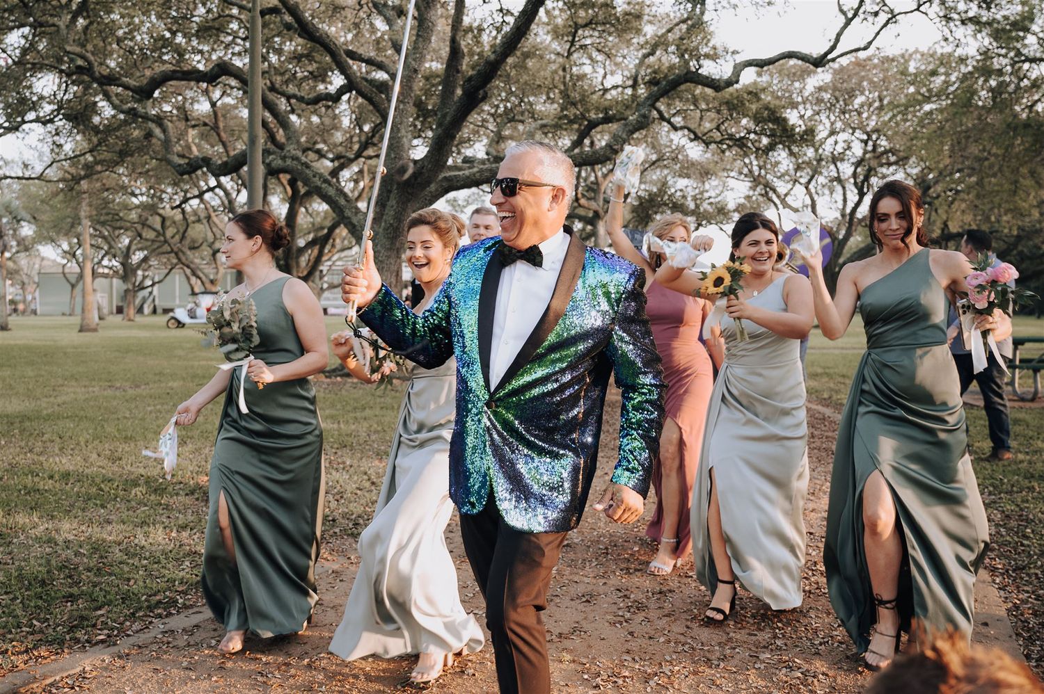 A wedding party follows a person in a sparkly jacket during an outdoor celebration.