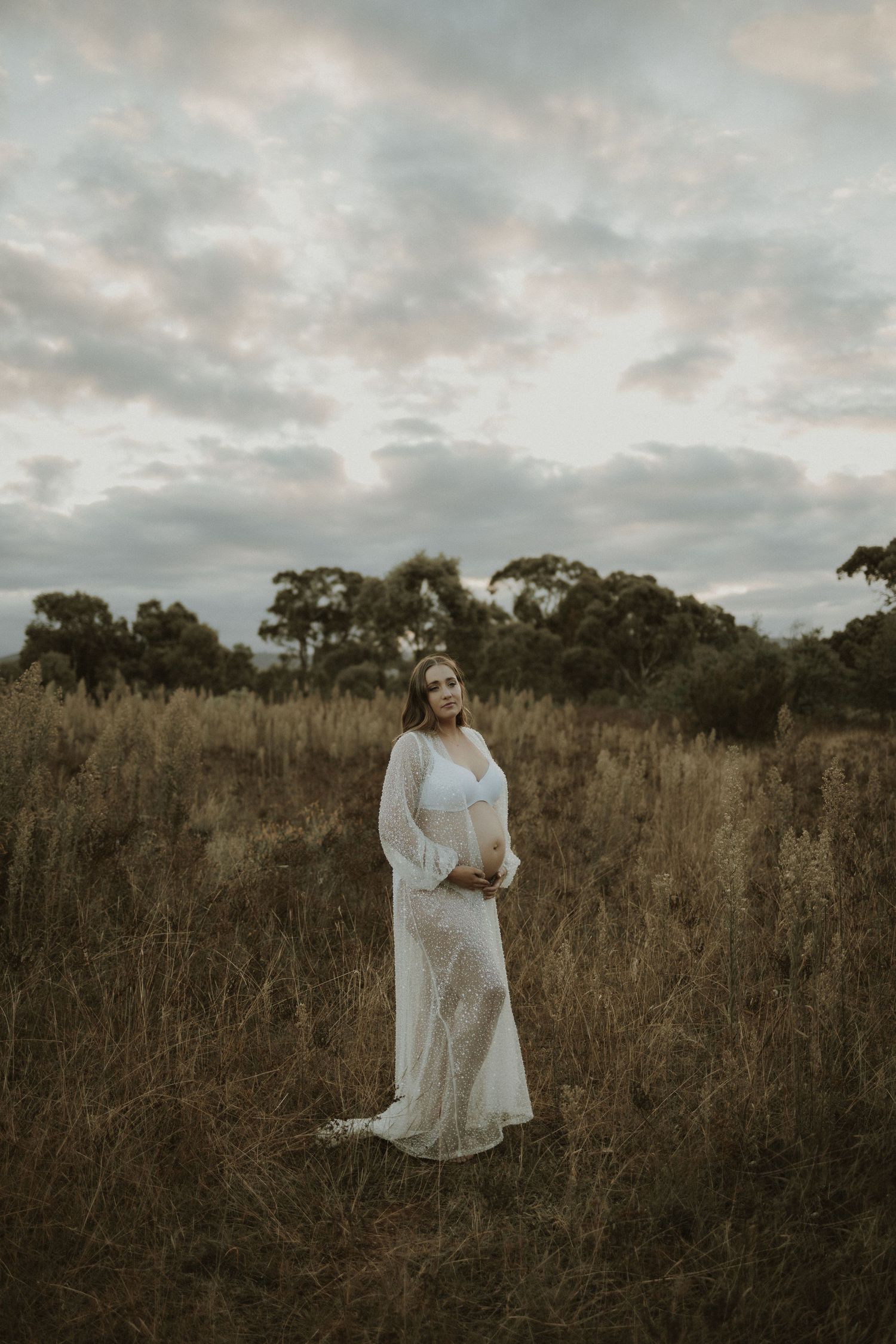 A woman in white lace poses in a field during sunset with dramatic clouds overhead.