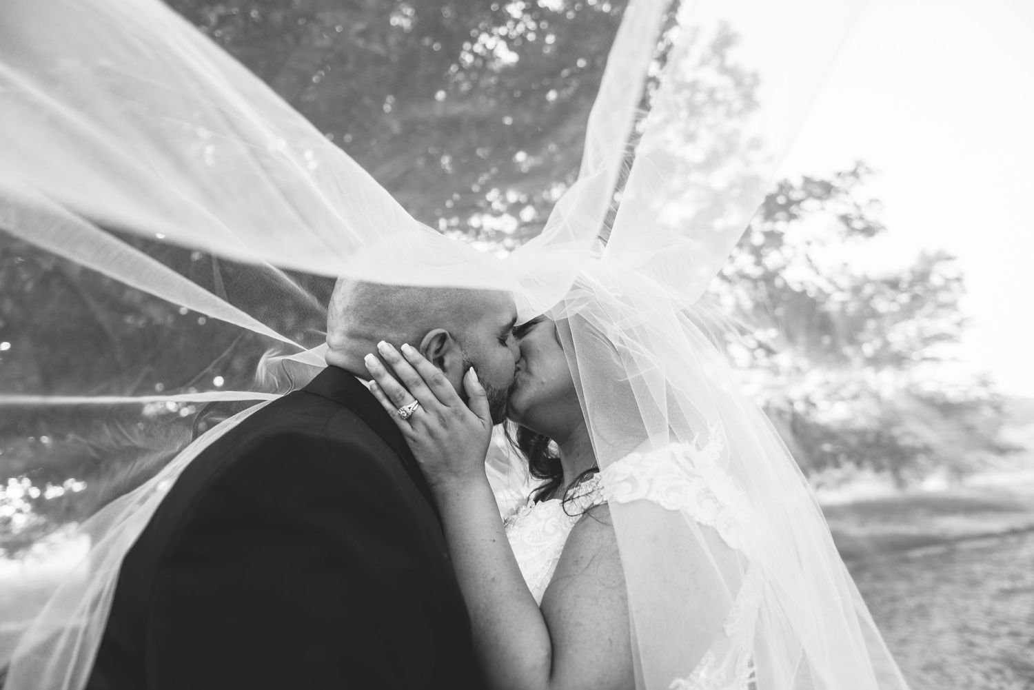 Bride and groom sharing a romantic kiss under a flowing veil in a black and white wedding photo.