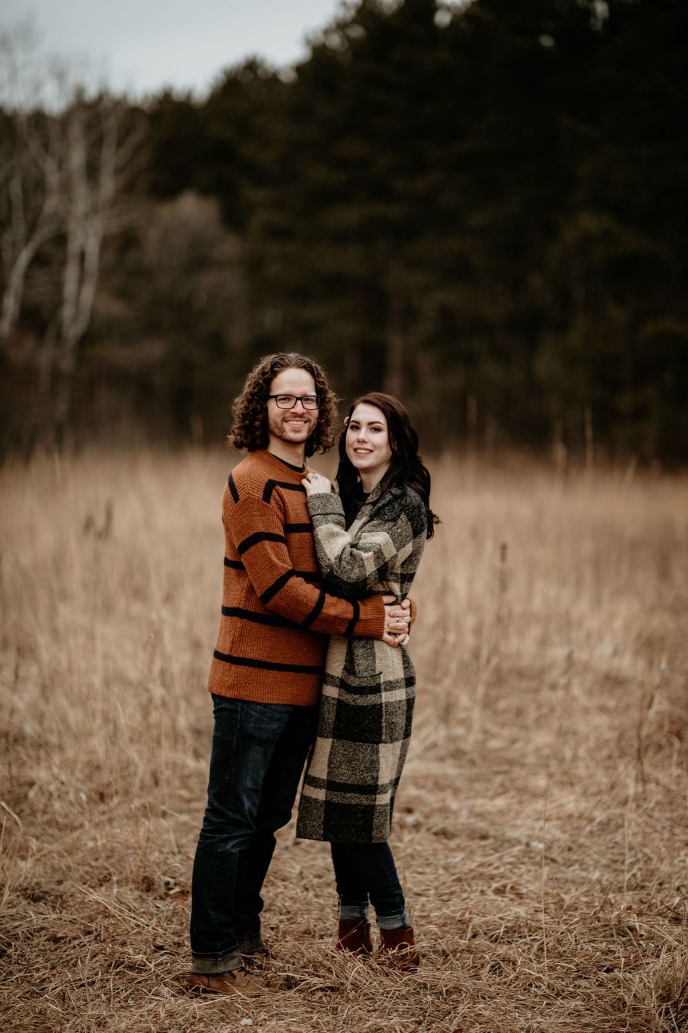 Smiling engaged couple embracing in golden autumn field, man in orange sweater, woman in plaid coat.