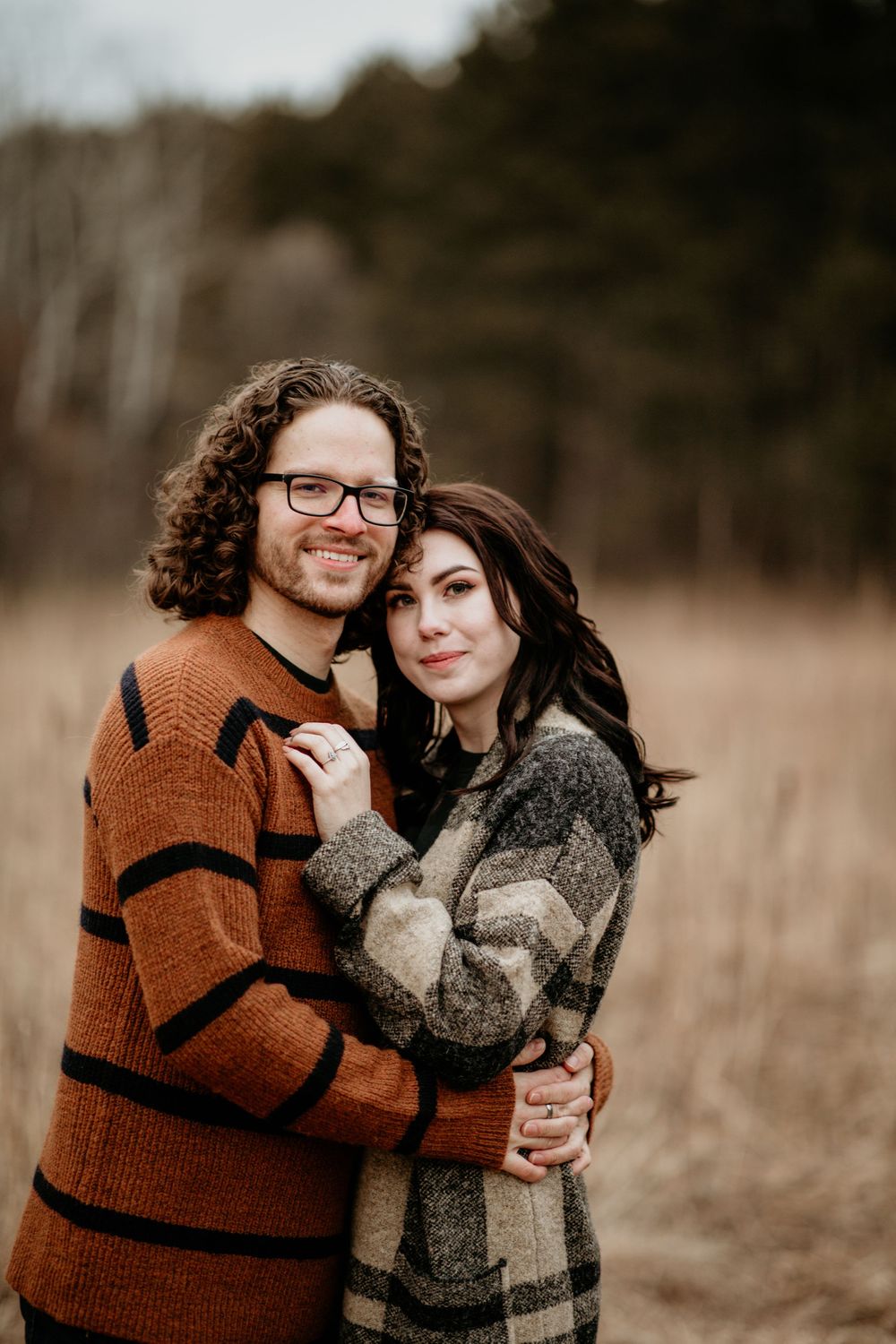Happy couple cuddling closely in dry autumn meadow, man in rust striped sweater, woman in plaid coat.