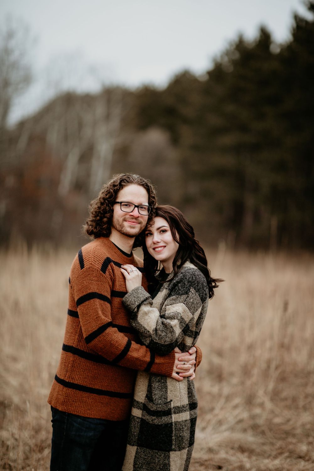 Joyful couple posing in winter meadow, man in orange striped sweater embracing woman in plaid coat.