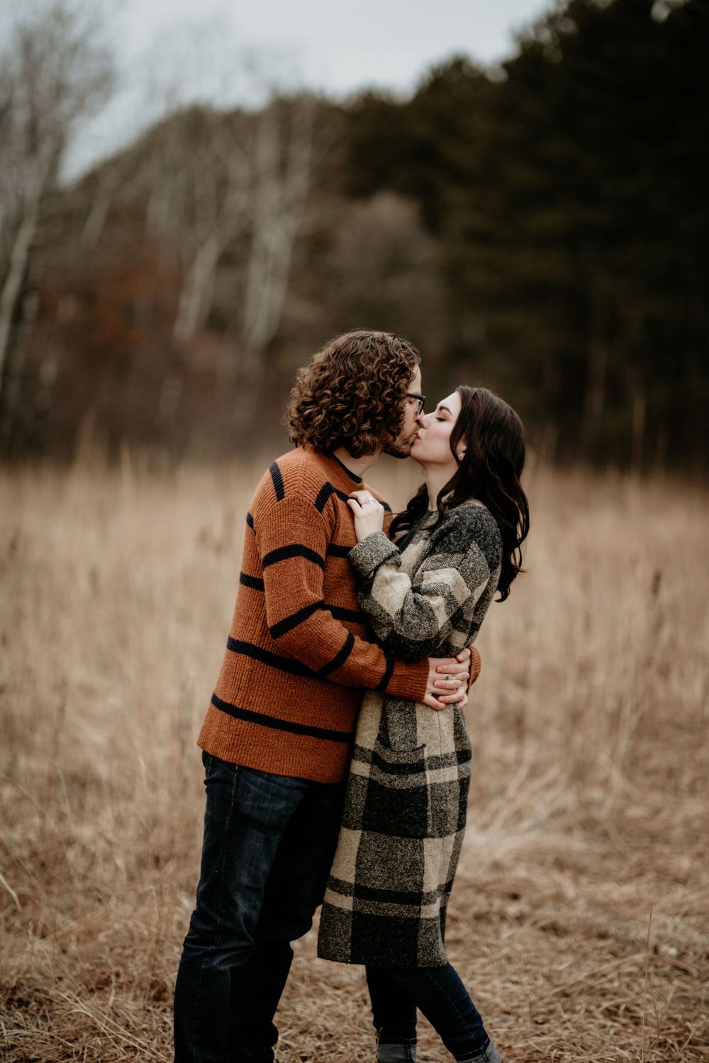Couple sharing a romantic kiss in golden fall field, wearing cozy sweaters and plaid outerwear.