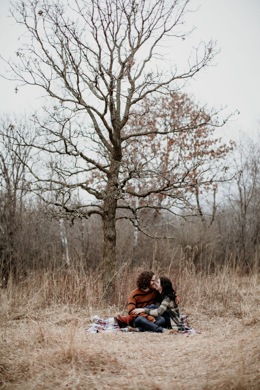 Two people sitting close together on a blanket under a leafless tree in a dry autumn meadow.