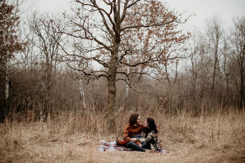 Couple sharing an intimate moment on a blanket surrounded by tall golden grass and bare trees.