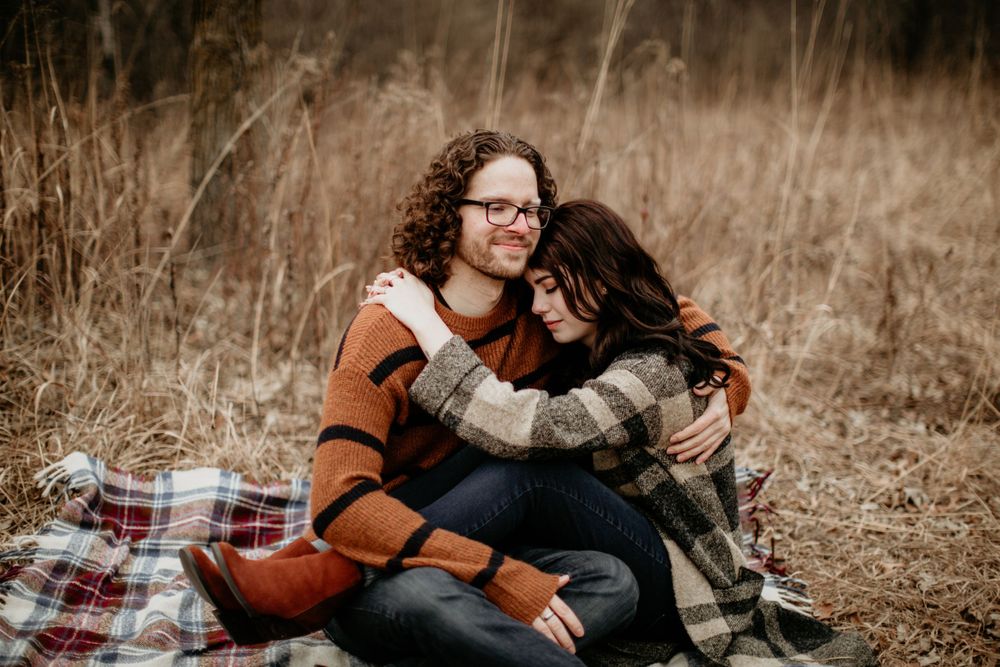 Smiling couple embracing on a plaid blanket, man in orange striped sweater hugging woman in plaid coat.