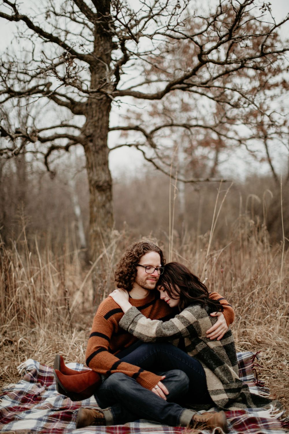 Couple embracing warmly on a plaid blanket beneath bare winter trees in a dry grassy field.