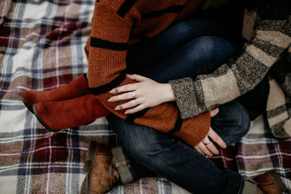 Close-up of couple holding hands on a plaid blanket, woman wearing engagement ring in autumn setting.