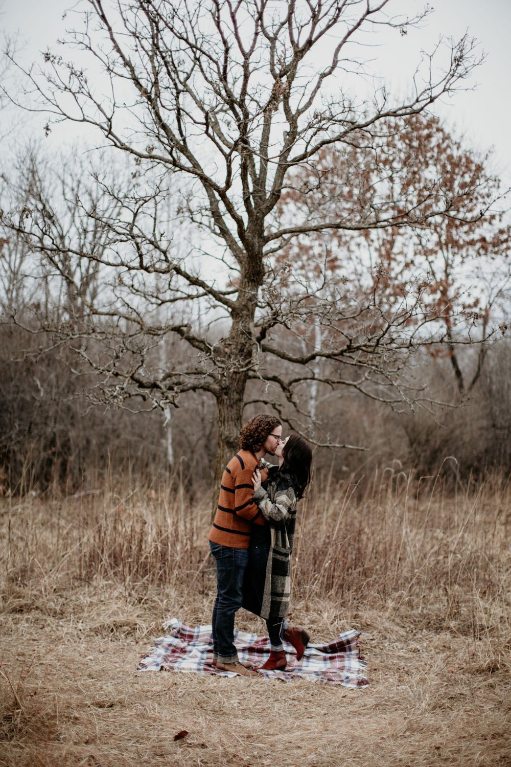 Couple standing and embracing on a plaid blanket beneath a large bare tree in a winter meadow.
