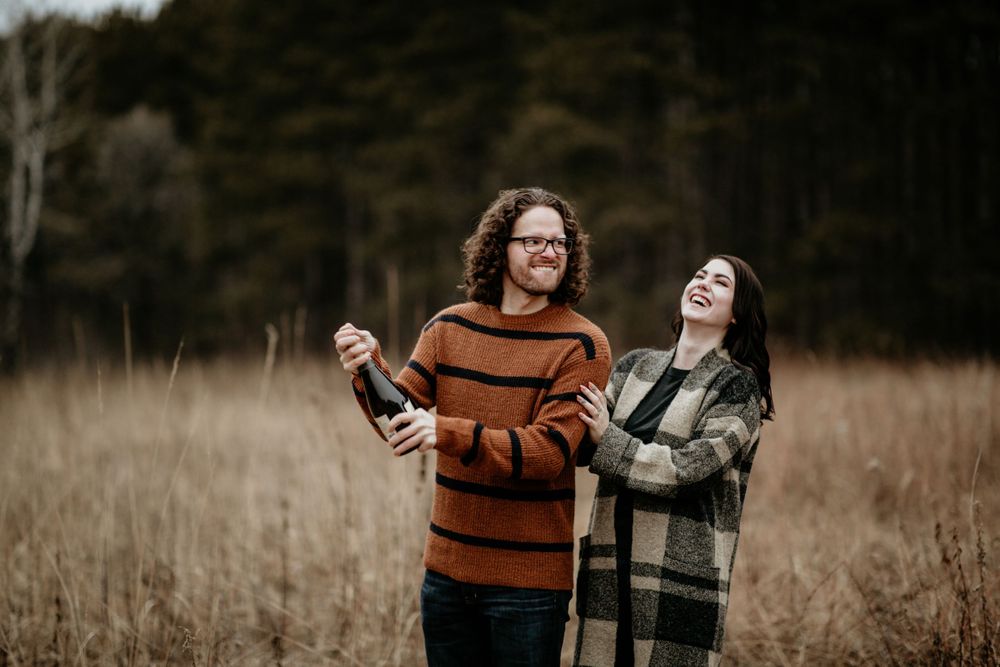 Couple laughing and opening champagne bottle in autumn field with brown grass and forest backdrop.