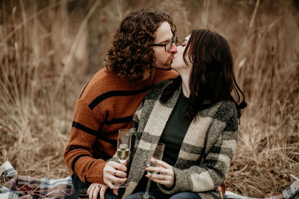 Couple sharing a romantic kiss while holding champagne glasses on a blanket in a fall meadow.