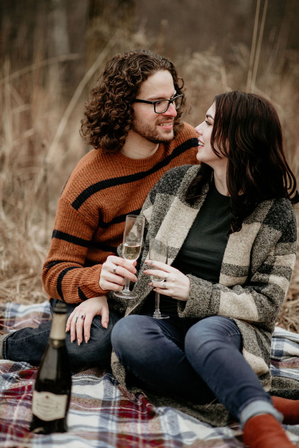 Smiling couple toasting with champagne glasses while sitting on a plaid blanket in autumn field.