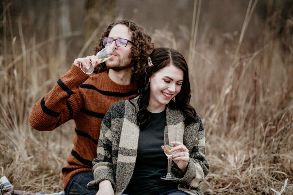 Couple sitting in tall dry grass, enjoying champagne together during an autumn outdoor picnic.