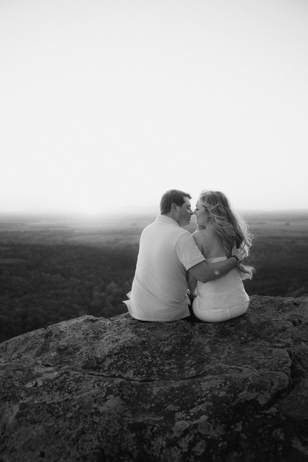 Black and white photography of a couple sitting on rocky terrain overlooking a vast landscape.