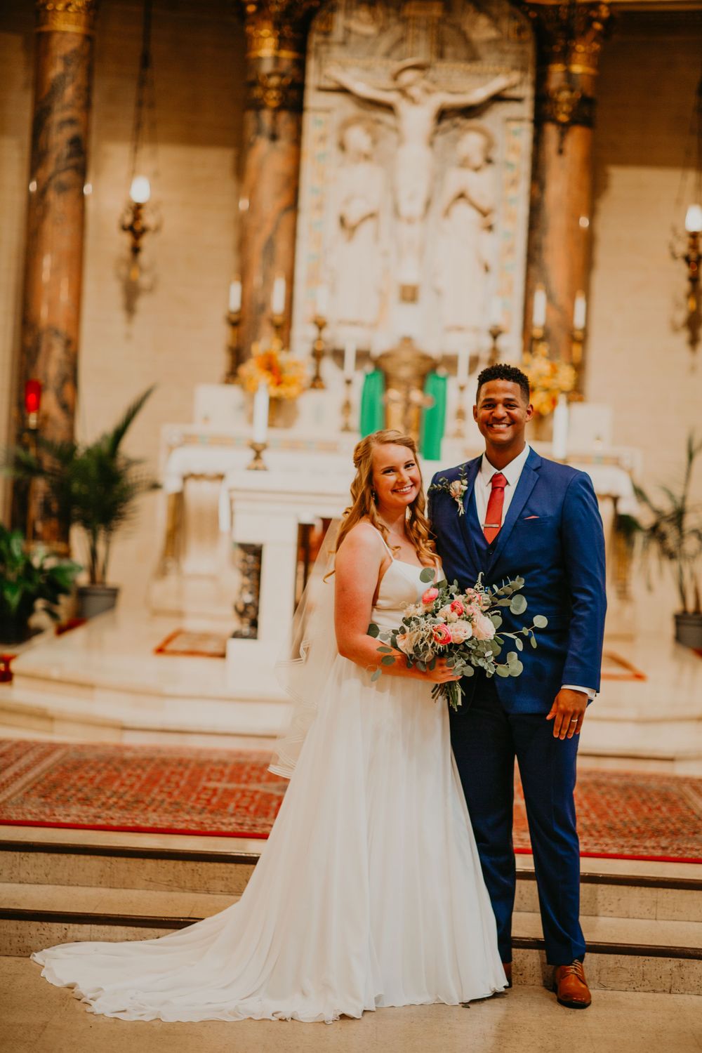 Happy newlyweds pose together in church, bride in white gown holding pink bouquet.