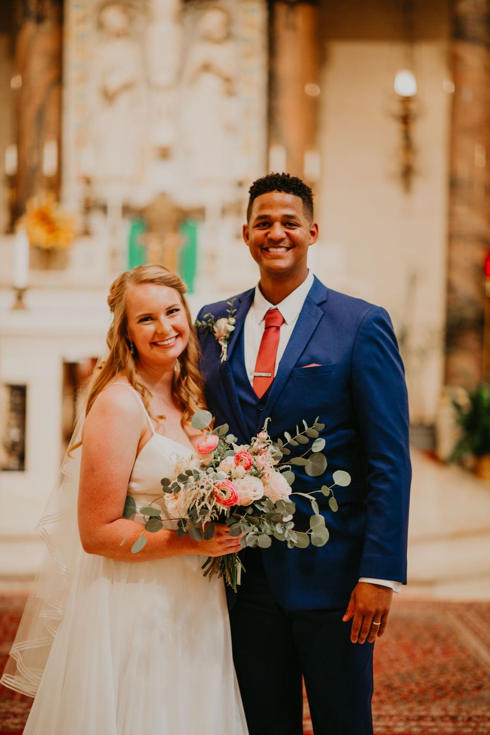 Smiling bride holds pink floral bouquet beside groom in blue suit inside church.