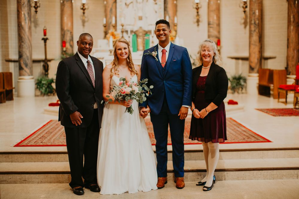 Newlywed couple smiles with two family members inside church following wedding ceremony.