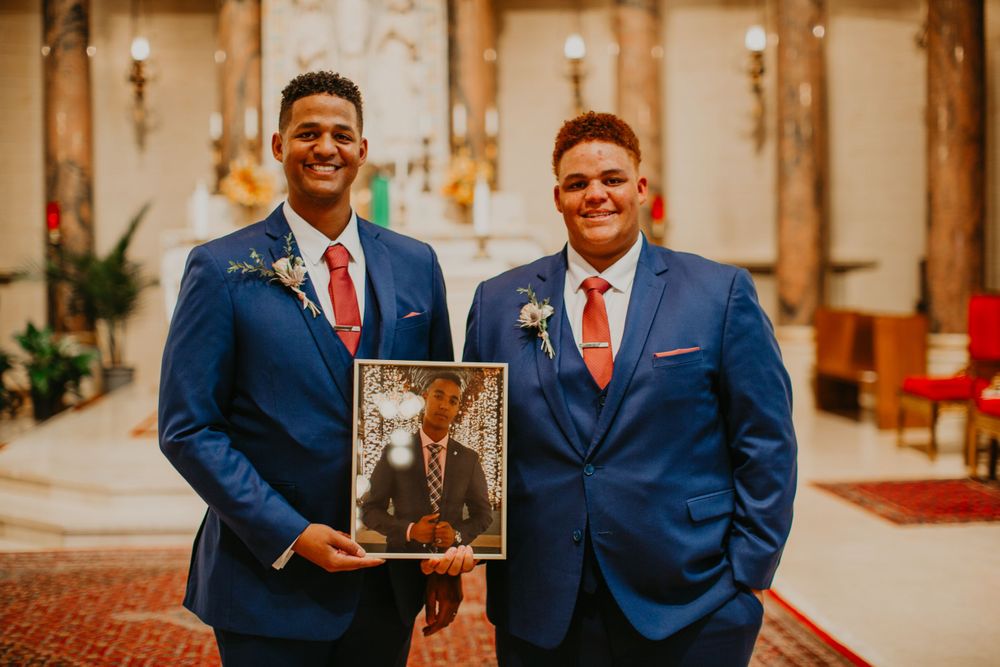Two men in blue suits pose with framed photo of loved one at church.