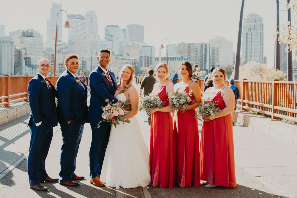 Wedding party poses on bridge with city skyline, bridesmaids in red gowns.