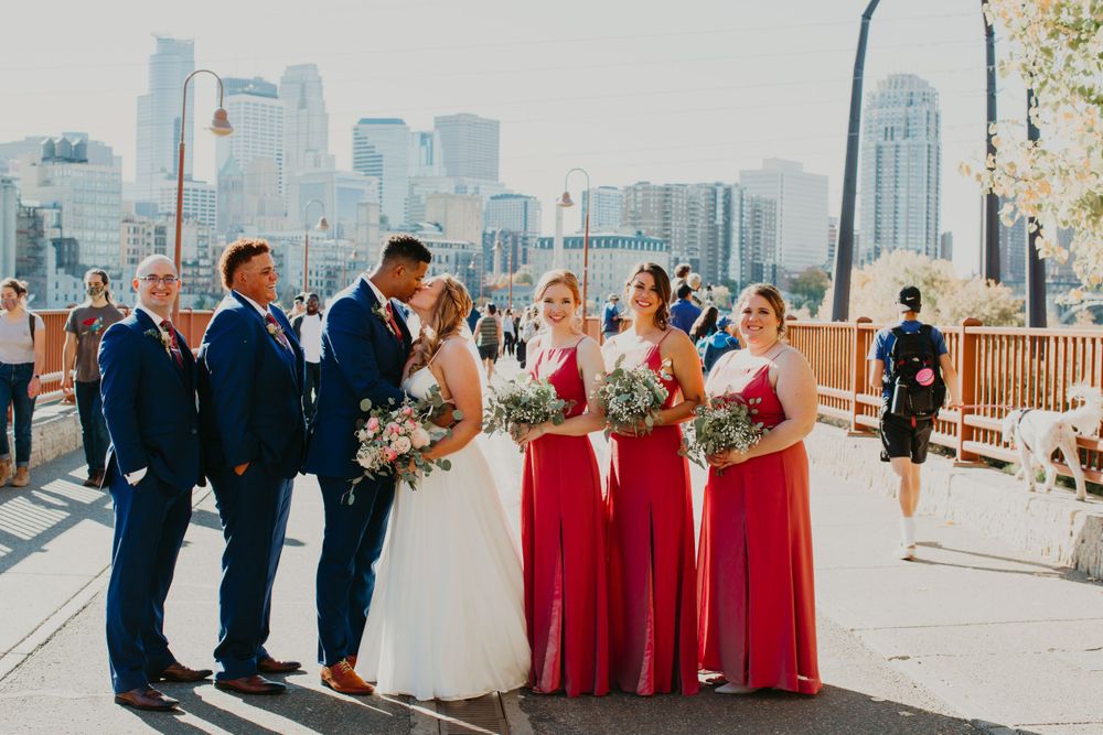 Wedding party poses on bridge with city skyline, bridesmaids in red gowns and groomsmen in navy suits.