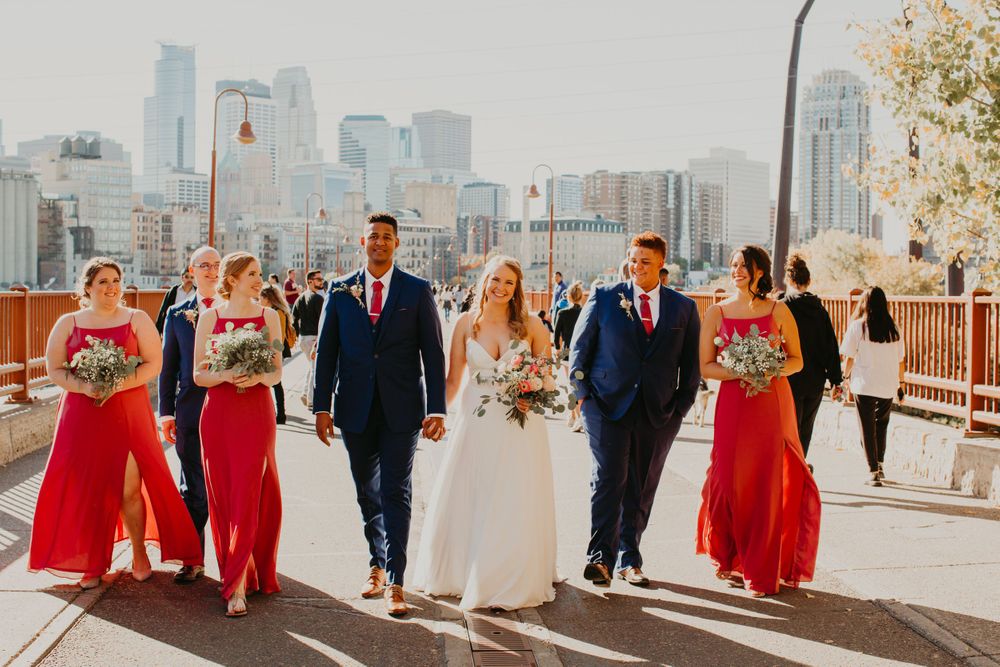 Wedding party walks on sunny bridge with city skyline, bride in white, bridesmaids in red dresses.