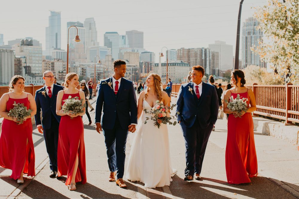 Bride and groom walk with wedding party on bridge, bridesmaids in red gowns, city skyline backdrop.