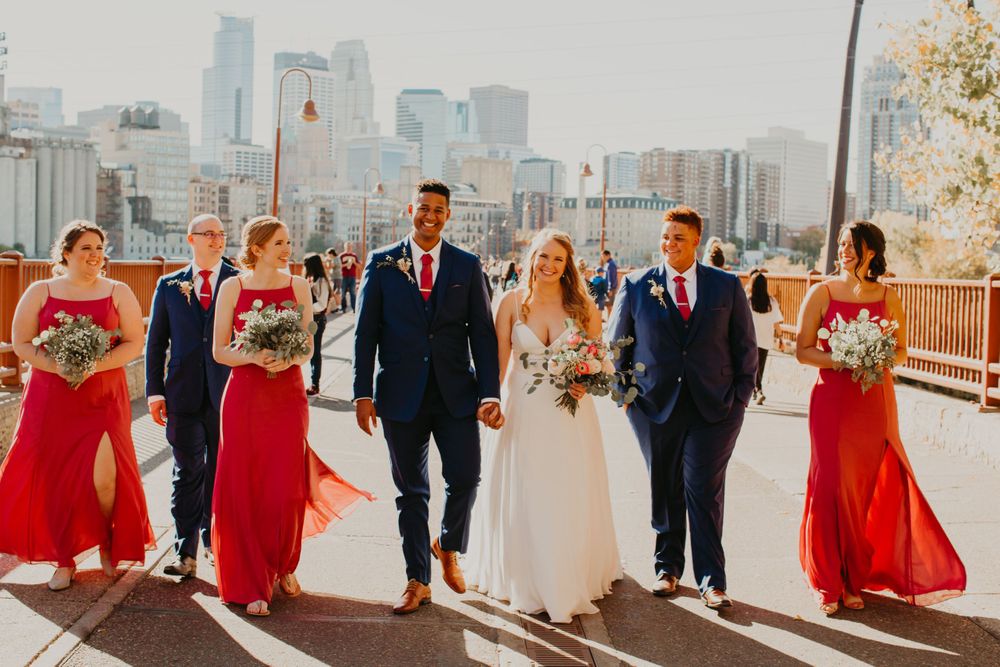 Smiling wedding party strolls on bridge with Minneapolis skyline, red bridesmaid dresses and navy suits.
