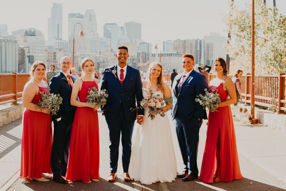 Wedding party poses with city skyline, bridesmaids holding bouquets in red gowns, groomsmen in navy.