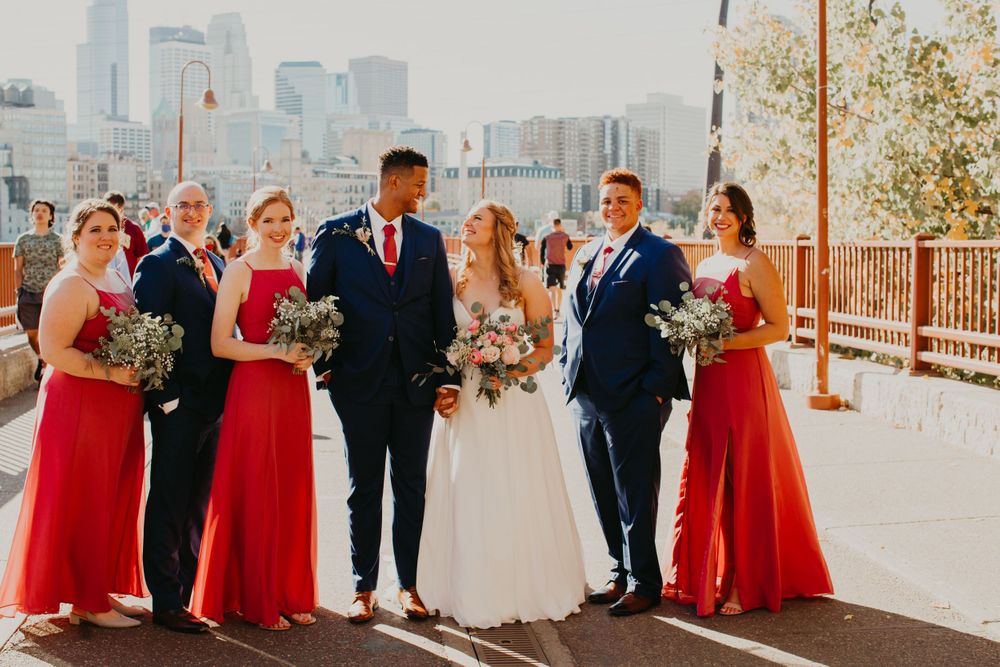 Wedding party stands together on sunny day with city skyline, red dresses and navy suits.