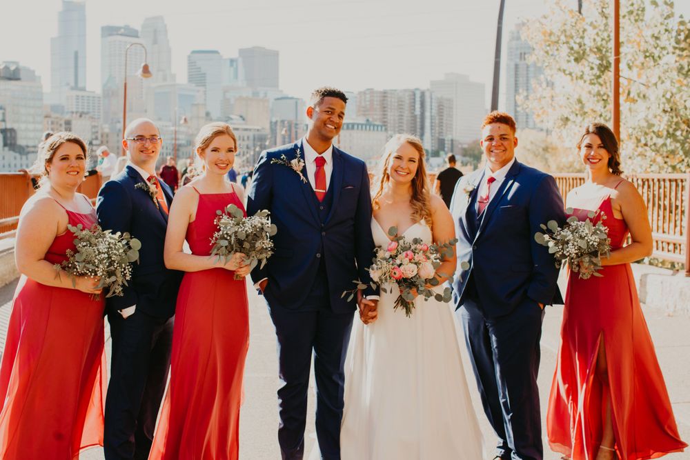 Laughing wedding party poses with Minneapolis skyline, bridesmaids in red gowns and groomsmen in navy.