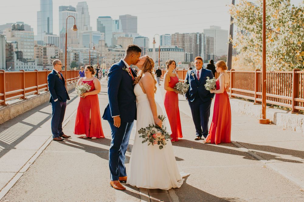 Bride and groom share a kiss on bridge with wedding party and city skyline in background.