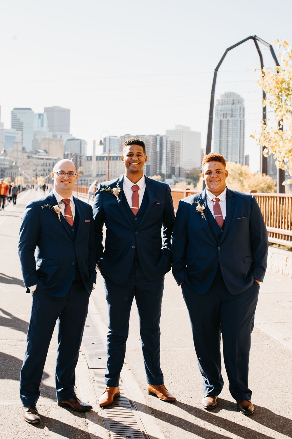Three groomsmen in navy suits with pink ties pose outdoors with city skyline background.