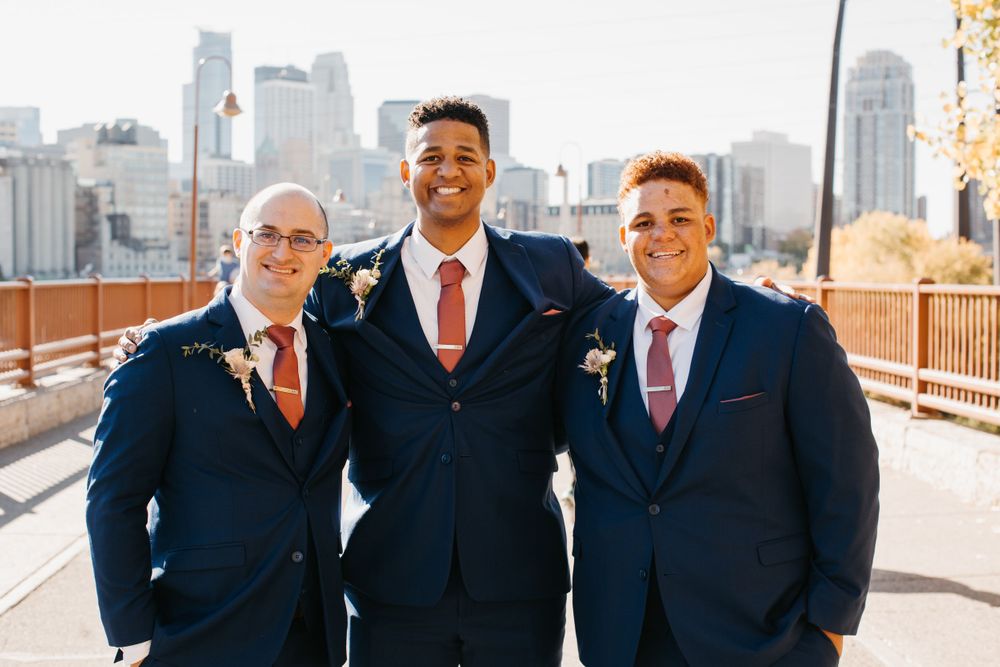 Three smiling groomsmen in navy suits pose together on bridge with city skyline behind them.