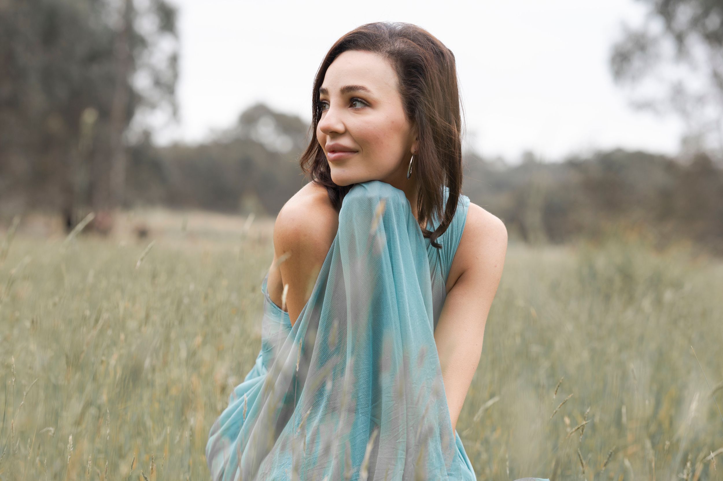 Woman in elegant teal backless dress looking over her shoulder in a scenic grassy meadow.