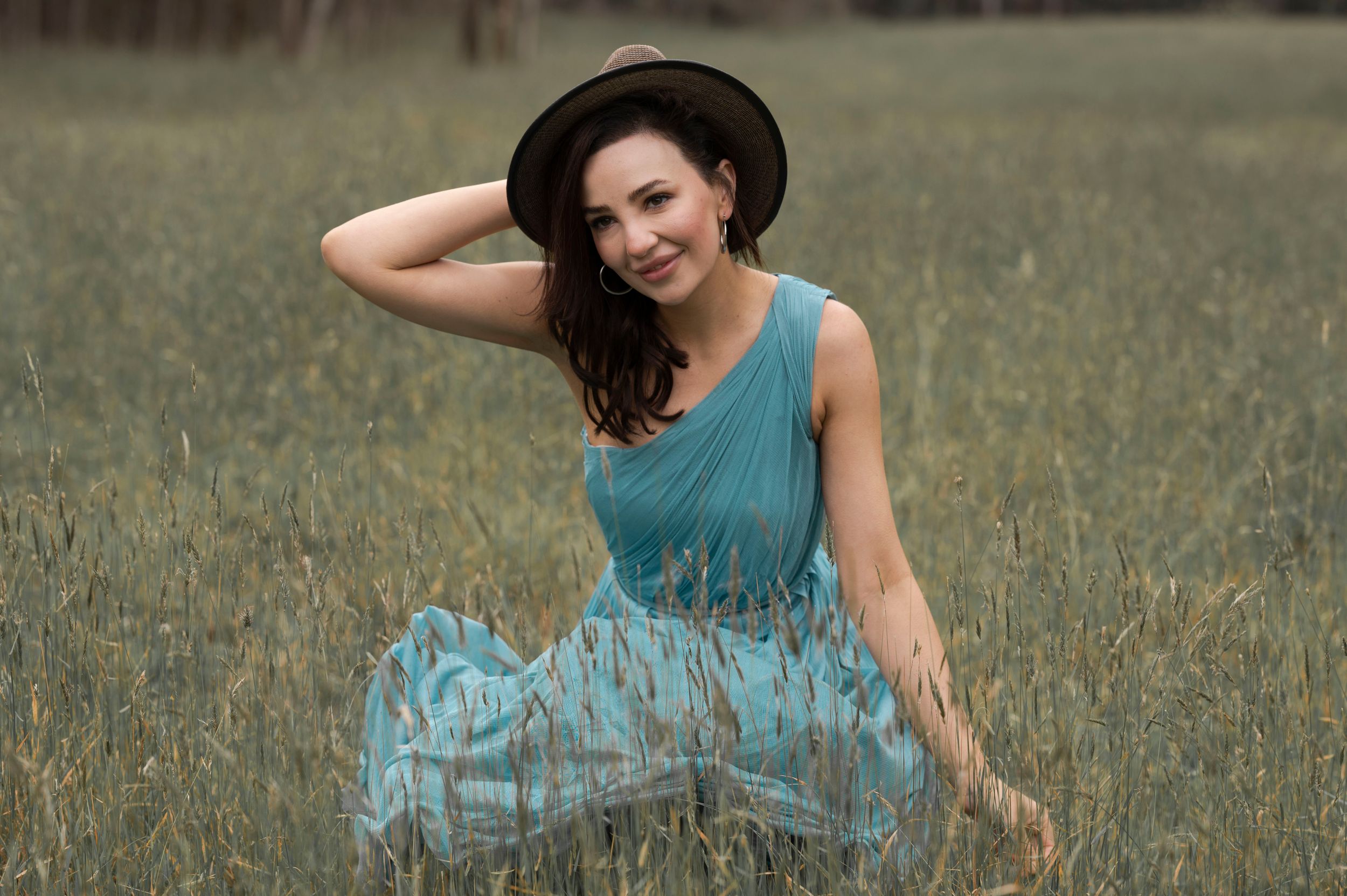 Smiling woman in teal dress and black hat sitting in a lush green grassy field.