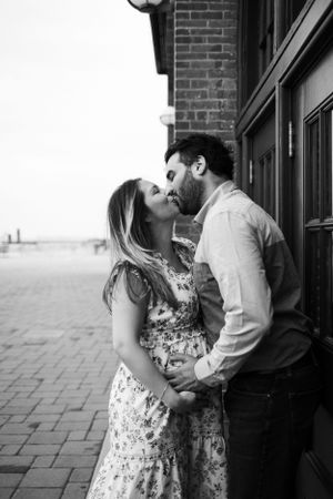 Intimate black and white portrait of expecting couple sharing a kiss against brick wall.
