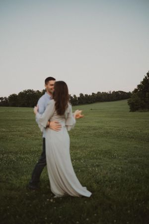 A couple embraces in a flowing white dress during golden hour in a meadow.