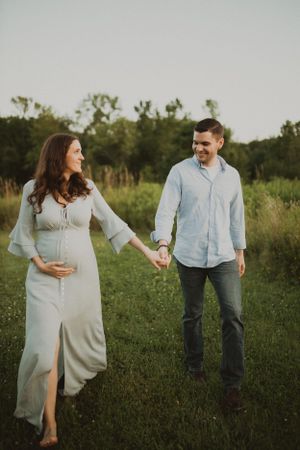 A pregnant woman in flowing white dress holds hands with partner in casual attire outdoors.