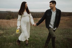 A couple walks hand in hand through tall grass, she in white dress and he in dark denim.