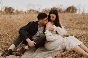 A couple sits together on a blanket in a golden field during a maternity photoshoot.