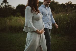 Expecting couple in light clothing share an intimate moment in a field at sunset.