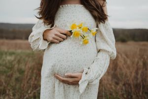 A pregnant woman in a white dress holds yellow daffodils in a rustic field setting.