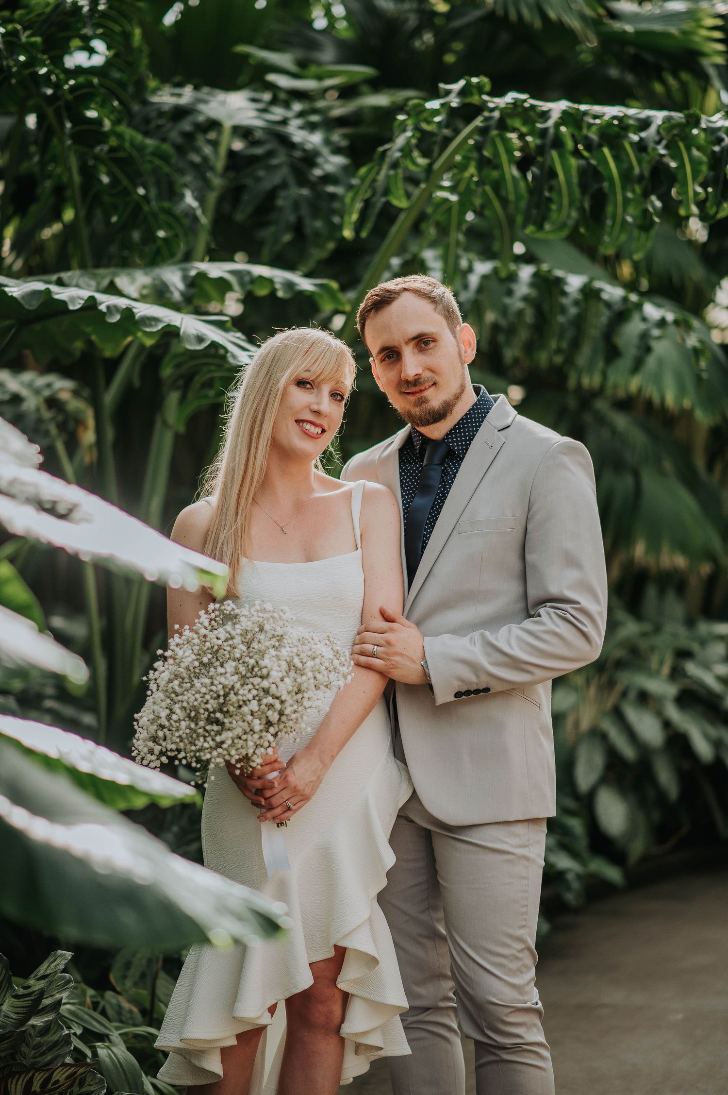 A couple poses with a bouquet against a backdrop of tropical greenery.