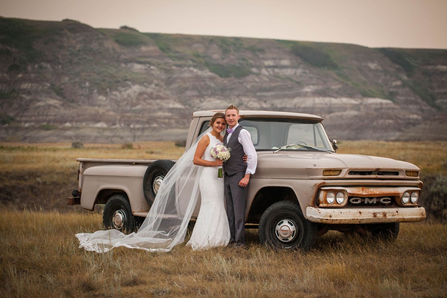 Newlyweds pose with vintage GMC pickup truck in rustic countryside setting.