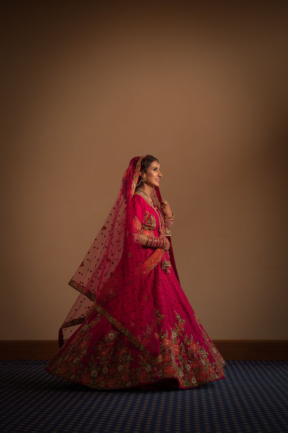 Traditional Indian bride poses in an ornate red lehenga with intricate gold embroidery against a brown backdrop.