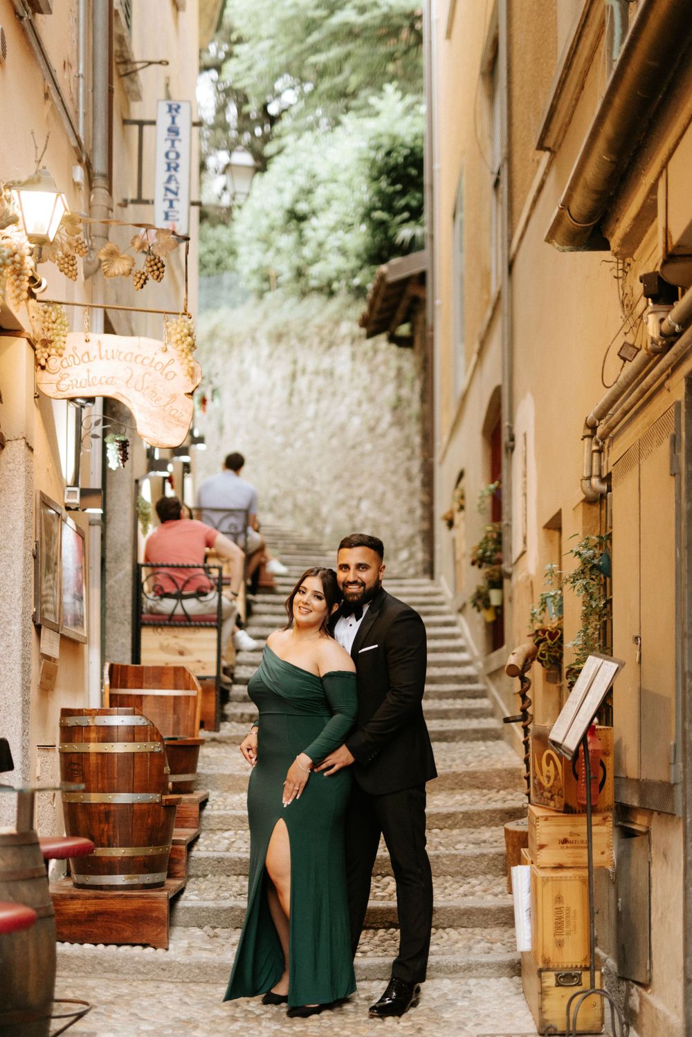 A couple poses romantically on charming cobblestone steps in a European alleyway with rustic barrels and lanterns.